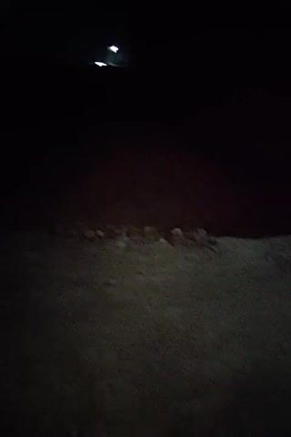 The video begins with a dark, blurry shot of what appears to be a ground surface, possibly dirt or gravel. The camera then pans up and to the right, revealing a dimly lit alleyway at night. The alley is narrow, with rough, concrete or plastered walls on either side. On the left, there is a dark doorway with a metal grate above it, suggesting a window or ventilation. Further down the alley on the left, another doorway is visible. The ground in the alley is uneven and appears to be made of dirt and debris. A single light source, possibly a streetlight or a light from a building, illuminates a portion of the alley from the right, casting long shadows. The overall impression is of a neglected and somewhat desolate urban environment.