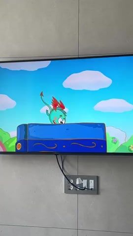 A television screen displays an animated scene. A pink pig-like cartoon character with a red hat stands in a grassy, green environment with trees and flowers. In front of the character is a closed, red-bound book. The character walks toward the book, then stops. The book opens, revealing pages with multiple images of cartoon faces and objects. The character then looks at the open book.