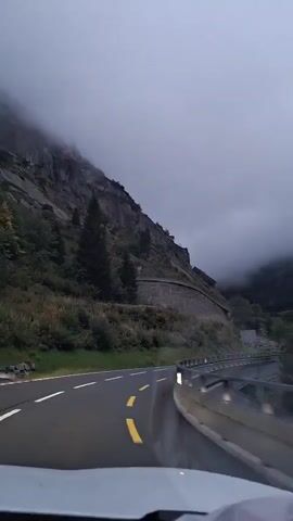 The video shows a view from the driver's seat of a car traveling on a winding mountain road. The road is wet and has yellow and white lane markings. On the left side of the road, there is a steep, rocky mountainside with some vegetation and trees. The mountainside is partly obscured by thick, gray clouds that hang low, suggesting foggy or overcast conditions. On the right side of the road, there is a metal guardrail. In the distance, several pairs of headlights from oncoming vehicles are visible, indicating the road continues into the fog. The overall atmosphere is muted and moody due to the weather conditions.