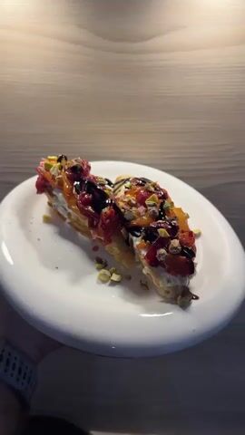 A white plate holds a dessert on a wooden table. The dessert appears to be a long pastry, possibly an eclair or a filled puff pastry. It is topped with a white cream, a reddish fruit compote or sauce, chopped pistachios, and a dark drizzle, likely chocolate. The dessert is cut in half, and the pieces are slightly separated. A person's hand, wearing a dark watch band, is holding the plate. The background is a light brown wooden surface. The lighting is warm and appears to be coming from above.