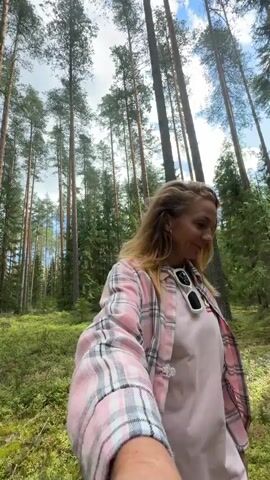 A woman with blonde hair, wearing a pink plaid shirt and a pink t-shirt, is walking in a forest. She is holding a camera in her right hand, as if recording a selfie video. She looks up at the sky, then turns her head to the side and smiles. In the background, there are tall trees and lush green vegetation. There is another person walking away in the distance. The camera is moving, suggesting the woman is walking.