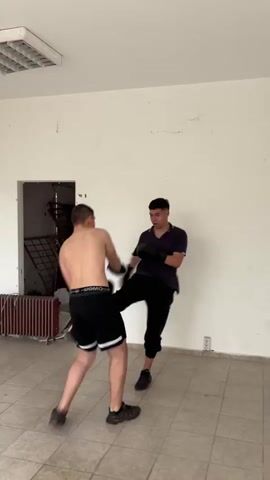 In a sparsely furnished, somewhat dilapidated room, two young men engage in boxing training. One is shirtless, wearing black shorts with white stripes and black athletic shoes; the other is dressed in a dark purple t-shirt, black pants, and black athletic shoes. Both are wearing black boxing gloves. The shirtless man throws punches at the gloved hands of the other, who appears to be using the gloves as training pads. The room has off-white walls with some visible wear and tear. A doorway with a view to an adjacent space, a radiator, and a ceiling light fixture are also visible in the scene. The floor is covered in light-colored ceramic tiles.