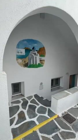 The video shows an outdoor area with a paved courtyard. The paving stones are irregular shapes with white grout between them. A white arched structure is visible on the left side of the frame, with a circular mural painted on its interior wall. The mural depicts a white building with a blue dome, a body of water, and green hills under a blue sky. The number "12-13" is painted on the upper left corner of the mural. In front of the arched structure, there are two small, rectangular windows. To the right of the archway, there is a low, white-plastered wall or bench. Beyond this, a series of small white tables with matching chairs are arranged along the wall, suggesting an outdoor seating area. Further to the right, a large white umbrella is open, with a table and chairs beneath it. The background consists of white buildings with blue accents, trees, and greenery. The scene appears to be in a Mediterranean setting, possibly a courtyard or patio of a cafe or guesthouse. The lighting suggests it is daytime, perhaps late afternoon or early evening, as the light is soft and diffuse. There are no people visible in the video.