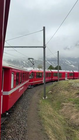 The video shows a long, bright red train rounding a bend on tracks through a green, grassy landscape towards snow-capped mountains veiled in mist. The perspective is from another train, looking out. The track bed is gravel, and power lines run above the train. The sky is overcast and grey. The number 37 is visible on a metal post beside the tracks.
