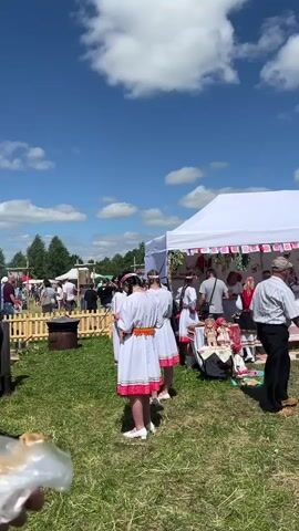 The video shows an outdoor festival on a sunny day with a blue sky and white clouds. In the foreground, a person is holding a piece of food, possibly bread. To the left, a wooden fence encloses an area with a metal barrel. In the center, a white tent is set up with decorations hanging from the front. Underneath the tent, several people are gathered, some wearing traditional white clothing with pink trim. They appear to be interacting with each other and possibly vendors. In front of the tent, there are tables displaying items that look like dolls and crafts. To the right of the tent, a group of people are standing and observing. Further in the background, more people are milling around, and trees form the horizon. A wooden signpost stands on the right side of the frame. The ground is covered in green grass.