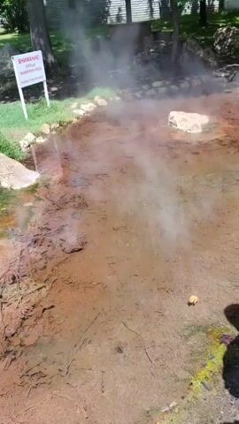 The video captures an outdoor scene featuring a natural hot spring in Karnalovo, Bulgaria.

The first shot (0:00-0:01) shows a wide, shallow pool of steaming, murky water in the foreground, indicating its high temperature. A white sign with red Cyrillic text is prominently displayed near the water's edge, reading: "ВНИМАНИЕ! ГОРЕЩА МИНЕРАЛНА ВОДА" (Attention! Hot Mineral Water). In the background, a woman wearing a white top and blue jeans sits on a rock under the shade of trees.

The second shot (0:01-0:02) provides a closer look at the edge of the water, showing tall green grass growing along the bank and a muddy, possibly algae-covered, bottom.

The third shot (0:02-0:03) pans across a large grassy area with several scattered rocks. In the distance, a verdant hillside with trees and a large, multi-tiered stone structure or ruins are visible under a clear, sunny sky.

The final and longest shot (0:03-0:06) shows a group of children and at least one adult standing on a dirt path next to the hot spring. The adult, dressed in dark clothing, holds a black bag or folder while facing the group. The children are dressed in various casual outfits, some with backpacks, and appear to be observing the steaming water. Lush green trees line the background, with a long fence or wall and a mountain range further in the distance. The steam from the hot spring is clearly visible, rising from the water next to the path where the group is gathered.

The overall scene suggests a visit to a natural attraction or park, possibly a school trip or guided tour, on a bright, sunny day.