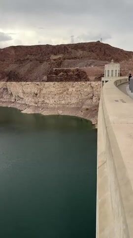 The video pans across a vast body of dark water, likely a lake or reservoir, with steep, rocky cliffs rising on either side. The cliffs exhibit distinct horizontal layers of tan and brown rock, indicating sedimentary formations. The water level appears to be significantly lower than the cliff face, revealing a watermark or a line of exposed rock. In the foreground, a portion of a concrete structure, possibly a dam or bridge, is visible, extending from the right side of the frame. The sky is overcast with gray clouds. The overall environment suggests a dry, arid region, likely a desert or semi-arid landscape. There are no people or animals visible in the video.