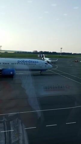 A Pobeda Airlines Boeing 737 aircraft is parked on the tarmac at an airport. The plane is white with blue accents and the Pobeda logo on the fuselage and tail. The reflection of another aircraft and airport markings can be seen on the surface in the foreground, indicating the shot is taken from inside another aircraft or a building with a reflective window. The sky is clear and bright, suggesting it is daytime.