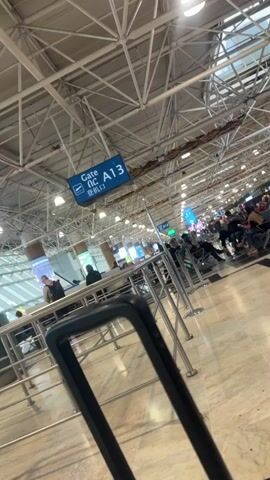 The video shows an airport gate area, presumably gate A13. The camera pans across the waiting area, revealing a large crowd of people seated and waiting. Many are looking at their phones or other devices. The gate is marked with a blue sign displaying "Gate A13" in English, French, and what appears to be an Asian language. The airport has a high, exposed metal ceiling with many lights. Several metal stanchions are visible in the foreground. The flooring is a light-colored, polished material. The overall environment suggests a busy airport terminal.