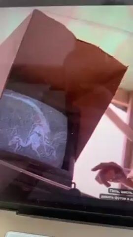 The video shows a person watching the movie Jurassic Park on a laptop. The laptop is on a wooden desk with a clear blue keyboard cover. The laptop is showing a scene where the characters are looking at a dinosaur skeleton on a small TV monitor covered with a cardboard sunshade. The person watching the movie touches the laptop screen near the TV image.