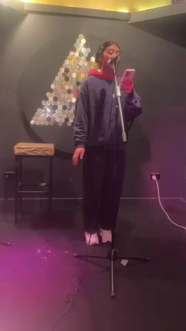A young woman stands in a dimly lit room, possibly a recording studio. She is holding a smartphone in her right hand and appears to be singing or speaking into a microphone positioned in front of her. She is wearing a blue hoodie over a red shirt, dark pants, and white sneakers. Behind her, a circular wall decoration with a triangular pattern of reflective discs is visible. To her left, a stool with a padded seat rests against the wall. The floor is dark and appears to have some pinkish lighting.