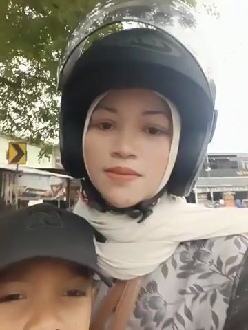 The video shows a woman wearing a black helmet and a light-colored hijab, riding on a motorcycle. She is looking at the camera and smiling. A child wearing a black baseball cap is visible in front of her, also looking towards the camera. The background appears to be an outdoor environment with trees and buildings, suggesting they are on a street. The video is a selfie-style shot from the perspective of someone on the motorcycle.