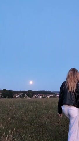 The video shows a young woman with long blonde hair from behind, wearing a black jacket and white pants, standing in a grassy field. The sky is clear and blue, with a full moon visible in the distance. In the background, there is a small town with houses and trees. The woman turns to face the camera and smiles, then spins around with her arms outstretched. She then turns back to face the moon and town, with her back to the camera.