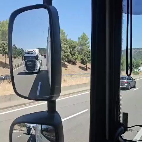 The video captures a first-person perspective from inside a truck cab, looking out at the highway through the side mirrors. The main mirror shows a white semi-truck with a white trailer in front, followed by other vehicles. The lower mirror reflects the interior of the truck cab. The scene outside is a sunny day with trees and hills in the background. The truck appears to be in motion on a highway, indicated by the blurry background and the movement of other vehicles.
