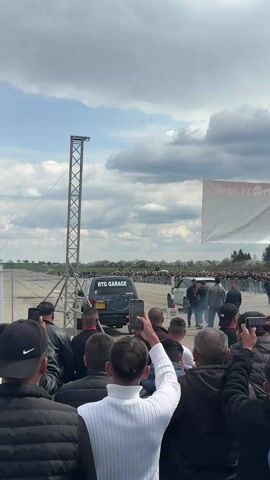 The video shows a crowd of people filming with their phones two SUVs preparing for a drag race. A dark blue Mitsubishi Pajero with “RTG GARAGE” printed in white on its back window is in the left lane, and a white SUV is in the right lane. Both cars are smoking their tires. There is a metal tower on the left of the frame, and a banner on the right. The background shows a flat field under a partly cloudy sky.