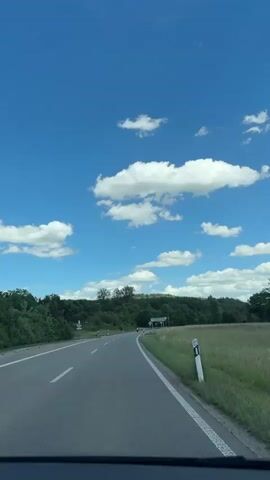 A motorcycle rider on a blue motorcycle with chrome accents is seen from a vehicle's perspective driving on a two-lane asphalt road on a sunny day. The road curves to the right. The sky is blue with white clouds. There are trees and grass on both sides of the road. There are road signs and white lines marking the lanes. The rider wears a helmet and dark clothing. The rider is moving away from the camera.