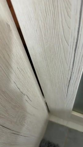 The video shows a gray and white cat peering out from a gap between two wooden-like surfaces. The cat has large, round, yellow eyes and its ears are slightly folded back. It appears to be looking upwards and to its left. The background is dimly lit and suggests an indoor setting with tile flooring visible at the bottom right of the frame.