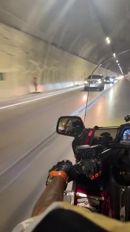 A first-person perspective video shows a person riding a motorcycle through a tunnel. The tunnel walls are concrete and illuminated by overhead lights. Cars are visible in the tunnel ahead, moving in the same direction as the motorcycle. The rider's gloved hands are on the handlebars, and their left arm with a tattoo is visible. The motorcycle is a red touring model with a windshield and mirrors. One mirror reflects a person wearing a yellow shirt. The video is shaky, indicating movement.
