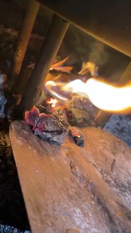 The video shows a close-up view of a fire burning on a shovel. Embers and flames are visible, with smoke rising from the fire. The shovel appears to be made of wood and metal. The background is dark and indistinct, suggesting an outdoor or dimly lit environment. The camera is positioned low, looking up at the fire and shovel. There are no people or other objects clearly visible in the frame. The fire is the primary focus of the video, with sparks occasionally flying from the burning material.