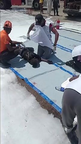 The video captures an outdoor emergency response drill or simulation, likely pertaining to a chemical spill or fire, given the extensive white foam covering the ground. The setting appears to be an industrial or transportation area, with "MAERSK" branded shipping containers visible in the distance.

Several individuals wearing white vests with "NIGERIAN RED CROSS" logos are actively participating, kneeling on light-colored tarps and administering first aid to "victims" lying on the ground. Some victims exhibit simulated injuries with fake blood. Other emergency personnel in orange shirts and yellow hard hats are also assisting. Red Cross first aid kits are visible nearby.

In the background, a red "FEDERAL FIRE SERVICE" truck with a contact number ("08032001557") and a white ambulance-style van, also bearing the Red Cross emblem, are present. The event is being filmed by at least two individuals operating video cameras on tripods.

Under a yellow canopy, a group of men, many in traditional attire, are seated, observing the proceedings. A banner behind them displays "NATIONAL EMERGENCY MANAGEMENT AGENCY (NEMA)" along with other organizational logos. The drill is taking place during a bright, sunny day.