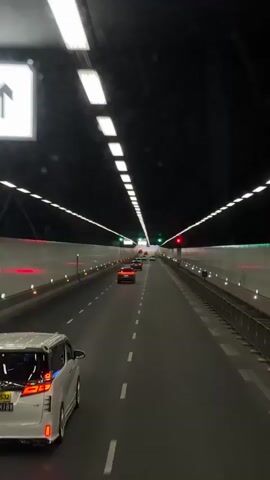 The video shows a view from inside a vehicle driving through a well-lit tunnel. The tunnel has a black ceiling with rows of bright, rectangular lights running along its length. Several vehicles are visible ahead, including a white van with a license plate in the foreground. The walls of the tunnel are white with evenly spaced red and green lights. The road is asphalt with white dashed lane markers. The general impression is of a modern, clean tunnel with moderate traffic.