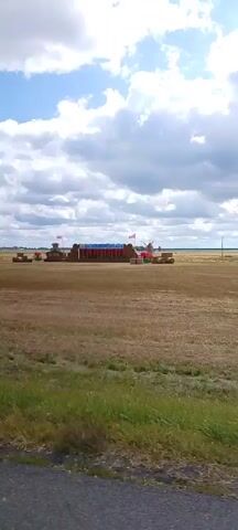 The video shows a wide shot of a harvested field under a cloudy sky. In the distance, a temporary stage or display is set up in the field. The stage appears to be made of hay bales and has flags and some structures on top. There are also some farm vehicles or machinery visible in the background. The camera pans slightly to the right, revealing more of the field and a roadside marker. The overall scene suggests an outdoor event or festival taking place in a rural setting.