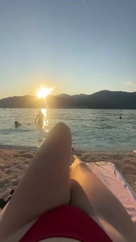 A first-person perspective shot captures a sunset on a beach in Donja Lastva, Montenegro. The sun, low on the horizon, casts a warm, golden glow across the water and the surrounding mountains, creating a serene and tranquil atmosphere. The sky is a clear, pale blue, with a faint contrail from an airplane visible.

In the foreground, the legs of a person are visible, lounging on a beach chair. The person is wearing a red bikini. A pink towel is draped over the chair, and a light-colored garment is folded on a nearby white side table. The beach itself is pebbled, with the gentle waves of the sea lapping at the shore.

In the middle ground, several people are enjoying the water. One person is partially submerged in the sea, their silhouette dark against the bright reflection of the sun. Another person is swimming further out. The water is clear and calm, with a slight ripple.

The overall scene evokes a sense of relaxation and peace, characteristic of a perfect summer evening by the sea. The combination of the natural beauty of the sunset, the calming sound of the waves, and the leisurely activity of beachgoers creates a picturesque and inviting setting. There is no visible text in the media file.