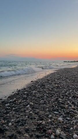 This is a vertical video of a beach at Nea Flogita, Greece, during sunset. The scene depicts a pebbly shore leading to a gentle sea with small waves. The sky transitions from a clear blue at the top to soft pink and orange hues near the horizon, indicating the time of day as sunset or twilight. The water appears calm with subtle ripples, and the pebbles on the beach are varied in size and color, ranging from dark gray to white and speckled. There are no visible people or distinct objects, with the exception of a faint distant light or structure on the far right, potentially a building or light source along the coastline. The overall mood is peaceful and serene, emphasizing the natural beauty of the coastal landscape at dusk. There is no visible text in the video.
