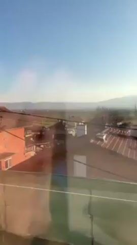 The video shows a view from a window or balcony, looking out over a landscape of houses and fields under a clear, bright sky. The sun is visible and causes glare on the glass. The landscape stretches to the horizon, where a distant mountain range can be seen. A green mesh barrier is partially visible in the foreground, attached to the railing of the balcony or window. The roofs of the houses are varied in color, with some appearing red-tiled. There is no apparent human presence visible within the landscape or interacting with the scene.