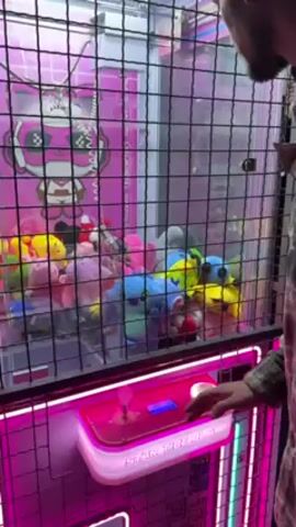 A man operates a claw machine. The machine is brightly lit with pink and white lights, and the control panel reads "STAR TIDE FUN". Inside the machine are numerous stuffed animals. The man uses the joystick to position the claw, then presses a button. He steps back as the claw lowers.