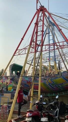 The video shows a carnival ride with a large, conical metal structure.  Two boat-shaped carriages, decorated with bright, colorful patterns, are suspended from the top of the structure by cables.  The carriages are in motion, swinging back and forth. Several people are visible in the carriages, some appearing to be passengers.  A person in a red shirt is standing on a metal ladder attached to the support structure, looking upwards.  In the foreground, several motorcycles are parked.  The sky is clear and appears to be daytime.  There are also colorful flags or banners strung across the top of the ride structure.