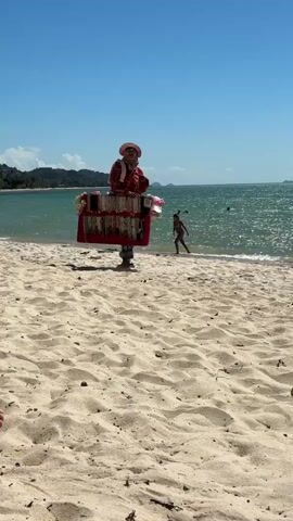 A person, dressed in a brightly colored outfit and a wide-brimmed hat, walks along a sandy beach. They are carrying a large, red display board laden with necklaces and other jewelry. The board is propped up by a strap around their shoulders. The person is walking towards the right side of the frame.

The setting is a beach with light-colored sand, leading to the ocean. The water is a vibrant turquoise and appears calm, with small waves gently lapping the shore. In the background, there is a coastline with lush green hills or mountains under a clear blue sky. The sun is shining brightly, casting a shadow from the person onto the sand, suggesting it is daytime. The overall atmosphere seems relaxed and tropical.

Further out in the water, a person is visible swimming or playing in the sea, and the outline of a boat or a distant island can be seen on the horizon. The presence of the jewelry vendor and the beachgoers indicates a tourist or recreational area. The scene evokes a feeling of a sunny, pleasant day at a seaside location. There is no visible text in the media.