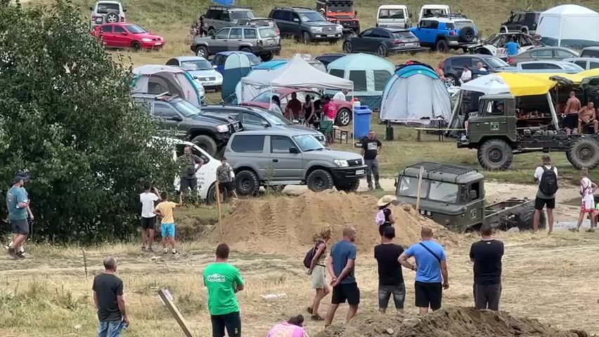The video shows an outdoor event, possibly an off-road vehicle competition or festival. In the foreground, several men stand watching. One man in a green shirt is facing away from the camera. To the left, a man in a black shirt is facing away from the camera. A group of people are gathered in the middle ground, some are standing and some are seated. A large, muddy military-style truck is driving over a dirt mound. Several other vehicles, including SUVs and cars, are parked in the background. Tents are set up in the background, suggesting camping or a gathering. A few people are walking around the parked vehicles. The environment appears to be a grassy, hilly area with dirt tracks.