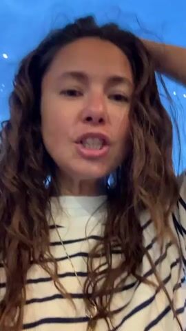 A young woman with long, wavy brown hair is looking directly at the camera. She is wearing a white t-shirt with horizontal navy blue stripes and a gold necklace. Her mouth is open, and she is speaking. She raises her right hand and touches her hair. The background is a blurry blue with some faint white lights.