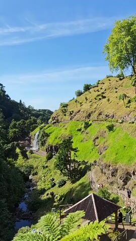 A vertical video shows a lush green mountainside with a waterfall cascading down a rocky cliff face. A stream flows through the valley below, and a pathway winds along the mountainside. There are several small buildings or structures visible near the waterfall and along the pathway, and a few people can be seen walking on the path. The sky above is blue with a few scattered clouds. The overall scene is one of natural beauty and tranquility.