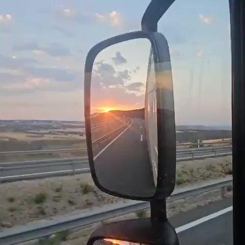 The video shows a reflection in a truck's side mirror of a sunset over a highway. The sun is low on the horizon, casting a warm orange glow across the sky and landscape. The highway curves through rolling hills, with a guardrail visible along the side. The reflection in the mirror shows the road stretching into the distance, with a few distant vehicles. The sky is a mix of blue and orange clouds. The video captures the beauty of the sunset and the open road.