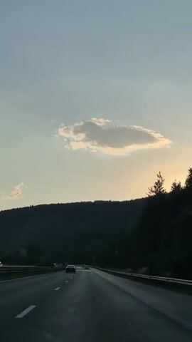 The video shows a view from inside a moving car on a highway. The scene is set in a hilly or mountainous area, with a dark, tree-covered hillside dominating the left side of the frame. A single, elongated cloud is visible in the pale blue sky, with a soft, golden light emanating from behind the hillside, suggesting it is either sunrise or sunset. The road ahead is clear, with a few cars visible in the distance. The overall atmosphere is serene and peaceful, with the gentle movement of the car creating a sense of travel. There is no visible text in the video.