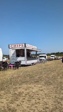 A white food trailer named "The Frying Squad" is parked in a grassy field under a clear blue sky. The trailer has a sign that says "CHIPS" on the side and "The FRYING SQUAD" on the front. The service window is open, and a person wearing a red shirt and a red hat is visible inside, preparing food. A menu board lists items such as "Bacon & Egg, Jumbo Hot Dogs, Burgers, Cheese Burgers, Hot Roast Pork, Bacon & Tom, Chips, Bacon Burgers." In front of the trailer, there are several plastic chairs and a small pink table. Behind the trailer, a white van is parked, and a line of other vehicles is visible in the distance. Several people are standing and sitting in the background. The ground is covered in dry grass.