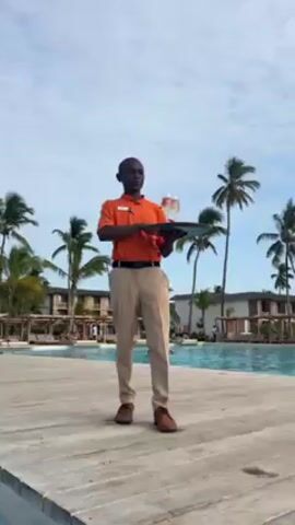 A short video shows a man working at a tropical resort. He is wearing an orange polo shirt and khaki pants, and he holds a tray with two drinks. He is standing on a wooden deck next to a large swimming pool. Beyond him are more palm trees and buildings. The camera then pans to a woman sitting in the pool, wearing a pink bikini and sunglasses. She smiles at the camera.