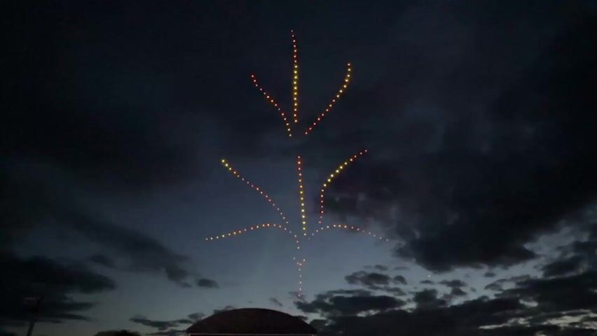 A video shows a dark sky with clouds at night. There are lights in the air that form a pattern resembling a tree.  The lights are yellow and spaced evenly. A structure appears silhouetted in the bottom of the frame. Music plays in the background.