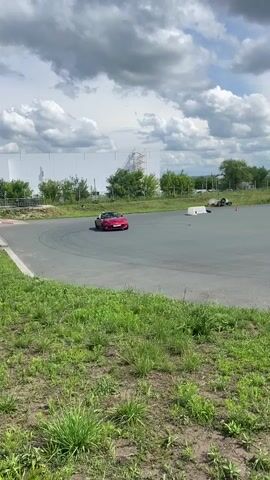 A red convertible Mazda Miata is shown driving on a paved track. The car has its top down and appears to have a driver and a passenger. The car is in motion, turning to the left. The track is surrounded by grassy areas and some trees in the background. There are orange traffic cones placed on the track, indicating a course or practice area. In the distance, there is a large white building and some other structures. The sky is partly cloudy.