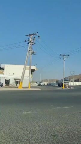 The video shows a roadside scene in what appears to be a desert or semi-arid environment. The sky is clear and blue. The initial shot features a gray minivan passing through an intersection. As the video progresses, various vehicles, including a white truck and several smaller trucks, pass by. On the side of the road, heavy construction equipment such as excavators and bulldozers are visible behind a fence. There is also a container-like structure selling water. Behind the road and vehicles, there are hills or small mountains. The camera seems to be positioned inside a moving vehicle, providing a driver’s perspective of the passing scenery. The overall impression is that the location is somewhat industrial or related to construction, with clear skies and a dry landscape.