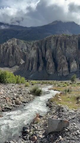 A wide shot of a rocky mountain valley with a narrow river flowing through it. The river is in the foreground, with rocks and sparse vegetation on its banks. In the mid-ground, a small line of trees can be seen to the left, and a single tree stands to the right. The background is dominated by a steep, rocky cliff face with numerous vertical formations. In the far background, a large, cloud-covered mountain peak looms. The sky is partly cloudy with dramatic lighting. The video is static, with no movement or people visible.