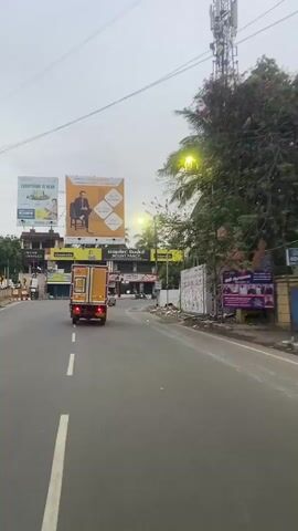This media file captures a street scene in St. Thomas Mount-Pallavaram Cantonment Board, India. The primary focus is a yellow and white box truck driving away from the camera on a grey asphalt road. The truck has red taillights and a license plate that reads "TN 20 B 4656". 

On the left side of the road, there are several large billboards. One billboard features a man in a suit and text that reads "WHY IS CASAGRAND THE FIRST CHOICE?". Another billboard has text in Tamil and English, including "MOUNT FANCY". Further down, there are shops with closed shutters. 

On the right side of the road, a building displays a sign with an eye logo and the text "EYEDEAL OPTICIANS". There are also street lights and electrical wires overhead. Trees with green foliage are visible, suggesting it might be daytime, possibly late afternoon or early morning given the overcast sky. The overall atmosphere appears to be that of a busy urban or suburban street. There are no people prominently featured, though there might be figures in the distance. The weather appears cloudy, but it's not actively raining. The scene is characterized by commercial signage and vehicular traffic.
