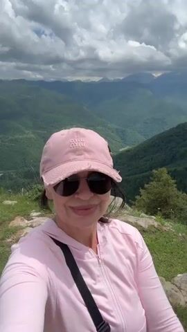 A woman in a pink baseball cap and sunglasses, wearing a pink zip-up hoodie, takes a selfie while standing on a grassy cliff overlooking a lush green valley with mountains in the background under a cloudy sky. In the background, two other women are visible. One woman is sitting on the ground looking at her phone, and another is standing further away. Later, a group of people is seen gathered on a grassy area in the distance, with tables and a red container.
