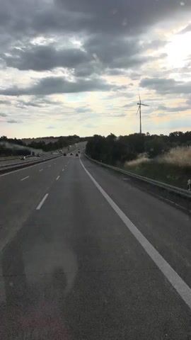 The video shows a view from a vehicle traveling on a highway. The highway has multiple lanes and is surrounded by trees and vegetation. There are other vehicles on the highway, and a windmill is visible in the distance. The sky is cloudy, with some sunlight breaking through. The overall scene suggests a daytime travel on a rural highway.