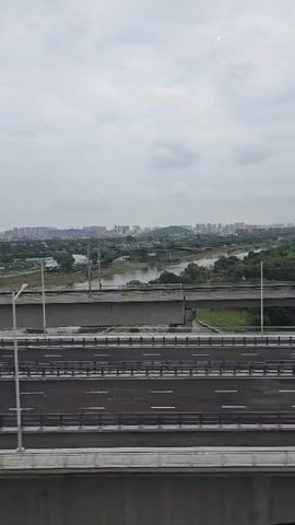 The video shows a view from a moving vehicle, looking out at a multi-level highway interchange. The sky is overcast, with a few faint bright spots, possibly from the sun peeking through clouds or reflections. In the foreground, there are at least three levels of highway visible, with concrete barriers and white lane markings. Several lampposts are also visible along the roadside. Beyond the highway, there is a body of water, possibly a river or lake, with lush green trees lining its banks. In the background, a dense urban landscape with numerous apartment buildings and other structures stretches towards the horizon. The buildings are mostly mid-rise, with some taller structures visible further in the distance. The overall impression is one of a modern urban environment with significant infrastructure development.