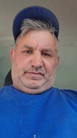 A medium close-up shot shows a man wearing a blue t-shirt and a blue baseball cap. His head is slightly tilted and he is looking directly at the camera with a serious expression. His hair is graying and messy, and he has a beard. The background is blurred and appears to be the interior of a vehicle. The man blinks and subtly shifts his gaze to the side before returning to the camera.