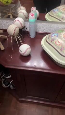 In a dimly lit room, a dark wooden dresser is covered in baseballs and softball. A mirror reflects the scene and a person's hands interacting with the objects. One person's hands are visible from the wrist down, wearing a dark shirt. Another person's hands, with visible rings and fingernails, are adjusting a yellow softball on a wooden handle, which is positioned like a tripod with a baseball on top. The softball is yellow and has black lettering. To the right of the softball, a collection of baseballs is arranged on the dresser. One baseball is on a metallic spider-like stand with multiple legs. Two other baseballs are clustered on top of the spider stand. Another baseball is placed on the floor in front of the dresser. To the far right of the dresser, a can of aerosol spray with a white and teal label is visible. In the reflection of the mirror, a group of people dressed in penguin costumes are seen standing in a line. The room appears to have shoes on shelves below the dresser.