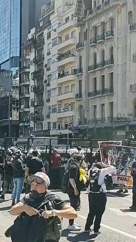 The video shows a street scene with a crowd of people standing near a line of police or security personnel behind barricades. Many of the people in the crowd are wearing masks or face coverings. Some individuals hold up signs or banners with images and text. Several buildings are visible in the background, including one with the sign "CAFEETERIA DEL MOLINO." The lighting is bright, suggesting a sunny day. The environment appears urban, with various architectural styles.